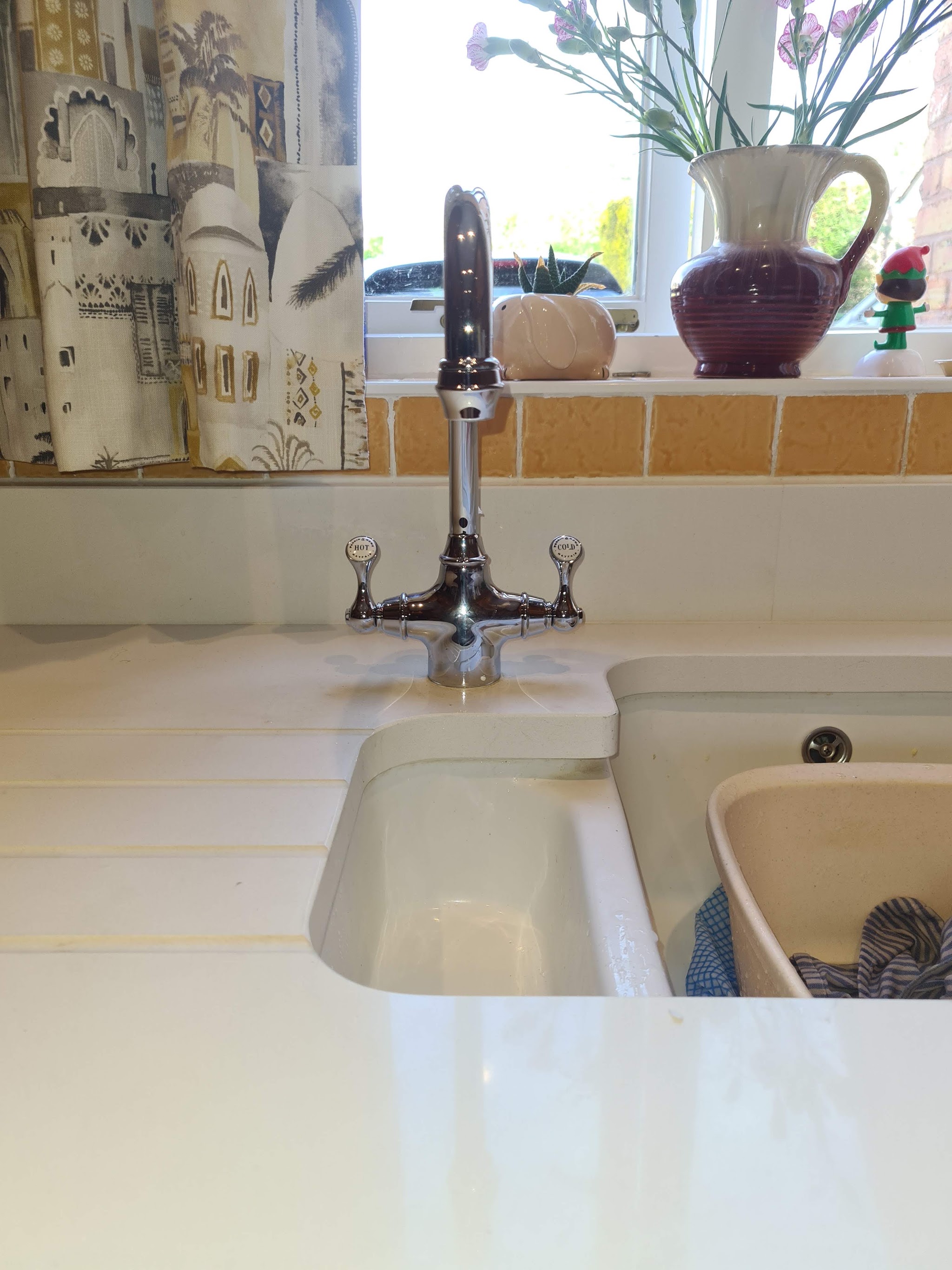A twin-lever chrome mixer tap over a white ceramic kitchen sink by a window